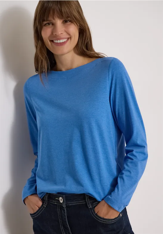 Frau im blauen Langarmshirt und Jeans, lächelt und steht entspannt mit Händen in den Hosentaschen vor hellem Hintergrund.