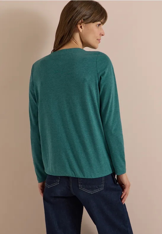 Frau von hinten in grüner Langarmbluse und blauen Jeans vor neutralem Hintergrund.