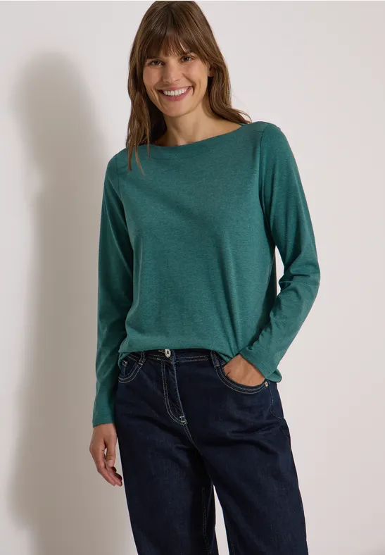 Eine lächelnde Frau in grünem Langarmshirt und blauen Jeans vor hellem Hintergrund.