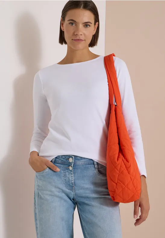 Frau in weißem Longsleeve und Jeans mit orangefarbener Stepp-Tasche vor hellem, neutralem Hintergrund.