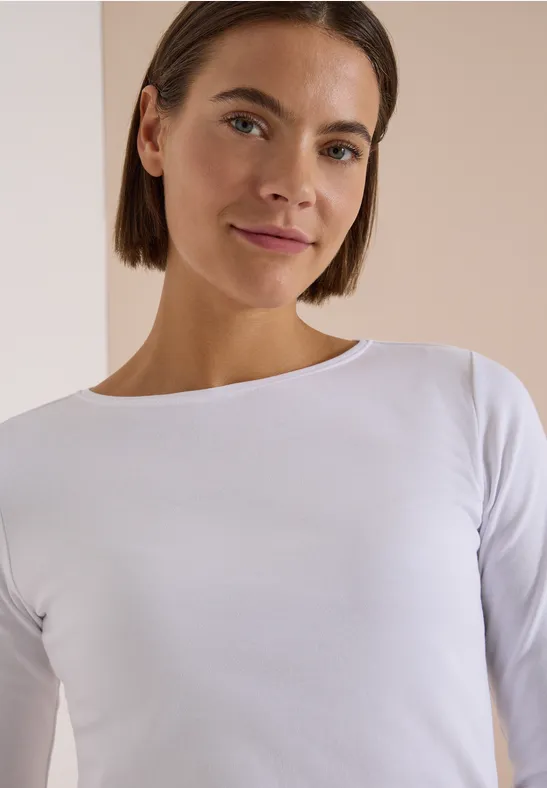 Junge Frau mit schulterlangem braunen Haar, weißem Shirt und leichtem Lächeln vor hellem Hintergrund.