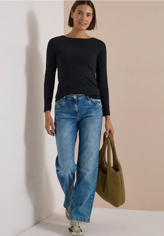 Junge Frau in schwarzem Langarmshirt und blauen Jeans mit grüner Tasche lehnt lächelnd an heller Wand.
