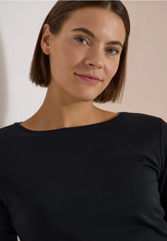 Frau mit kurzen braunen Haaren in schwarzem Langarmshirt, freundlicher Blick, neutraler Hintergrund.