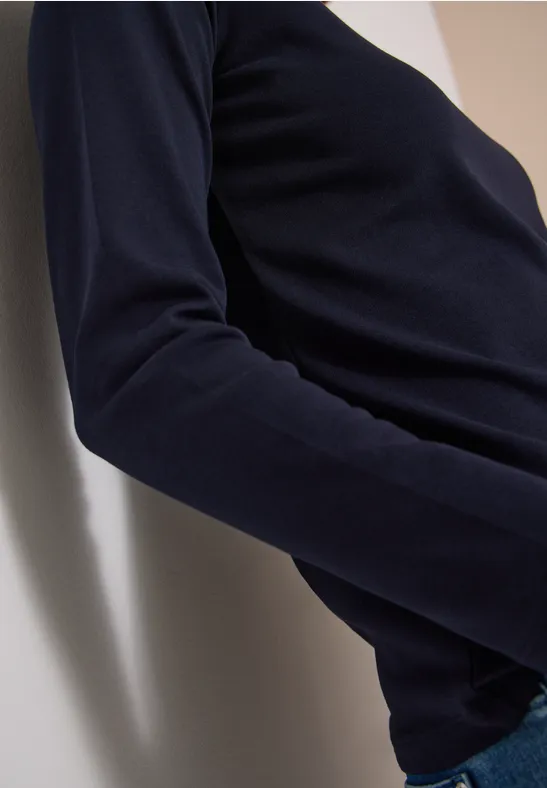 Person lehnt an weißer Wand, trägt dunkelblauen Pullover und Jeans