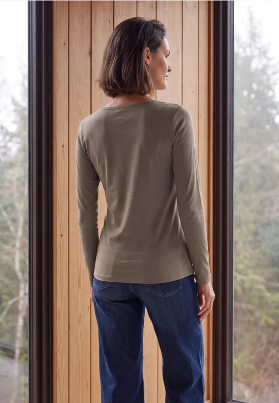Eine Frau steht vor einem Fenster, Rückenansicht trägt ein olivgrünes Langarmshirt und blaue Jeans.