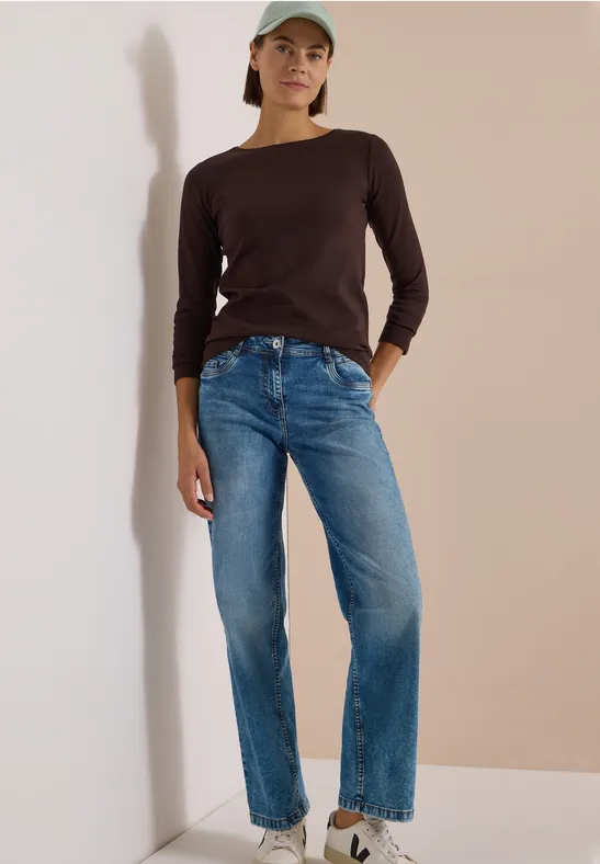 Frau lehnt lässig in blauen Jeans, schwarzem Longsleeve, Sneakern und Cap an hellem Hintergrund.