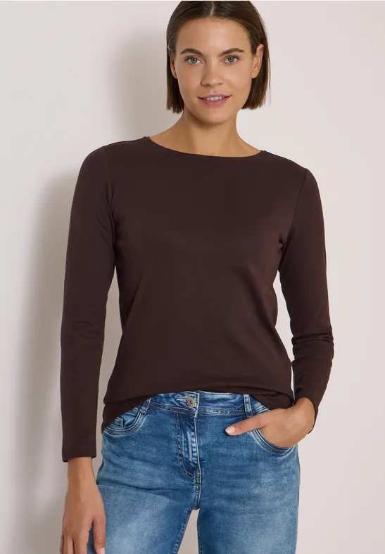 Frau mit braunem Langarmshirt und blauen Jeans, eine Hand in der Tasche, lächelnd vor neutralem Hintergrund.