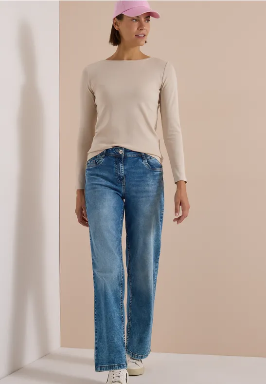 Eine junge Frau mit rosa Cap, beigem Langarmshirt, weiten blauen Jeans und weißen Sneakern lehnt lässig an einer Wand.