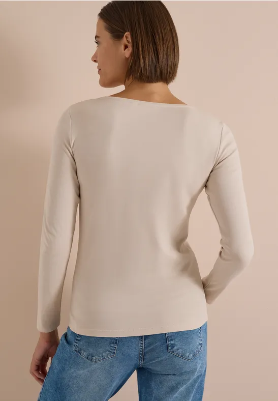 Frau von hinten in hellbeigem Langarmshirt und blauen Jeans vor hellem Hintergrund.