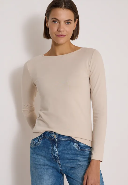 Eine Frau trägt ein beigefarbenes Langarmshirt und blaue Jeans vor weißem Hintergrund.