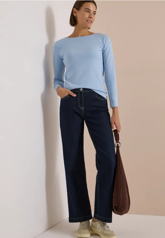 Frau steht mit hellblauem Langarmshirt, weiten dunklen Jeans und brauner Tasche vor einer Wand.