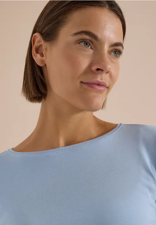 Nahaufnahme einer Frau mit kurzem braunem Haar und hellblauem Top vor neutralem Hintergrund.
