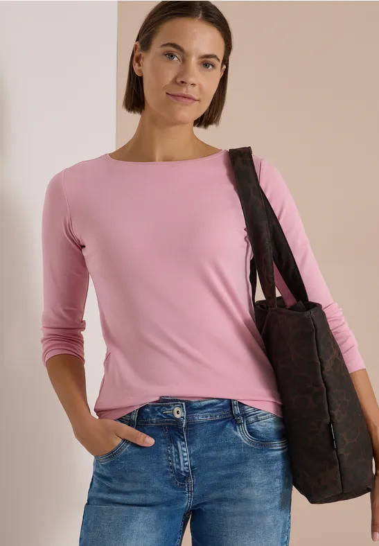 Frau trägt rosa Langarmshirt und Jeans, mit einer Tasche über der Schulter vor neutralem Hintergrund.