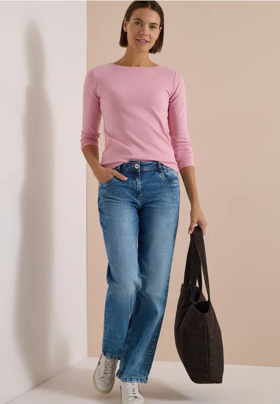 Eine Frau in rosa Longsleeve, blauen Jeans und weißen Sneakers trägt vor schlichter Wand eine schwarze Tasche.