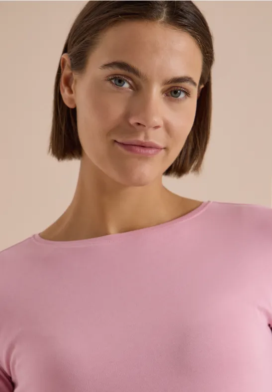 Junge Frau mit kurzen braunen Haaren und rosa Oberteil vor hellem, neutralem Hintergrund, leichtes Lächeln.