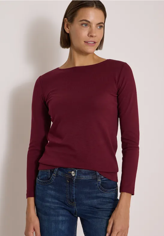 Eine Frau trägt einen dunkelroten Pullover und blaue Jeans, steht vor einer hellen Wand.