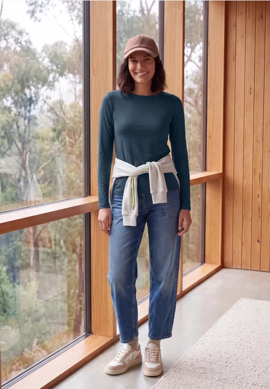 Frau in Jeans, grünem Pullover, Cap und Sneakers steht in hellem Holzraum am Fenster.