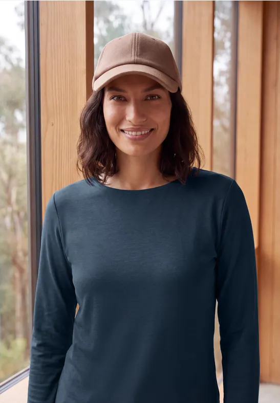 Eine lächelnde Person in einem dunkelblauen Pullover und Schirmmütze vor großen Holzfenstern.