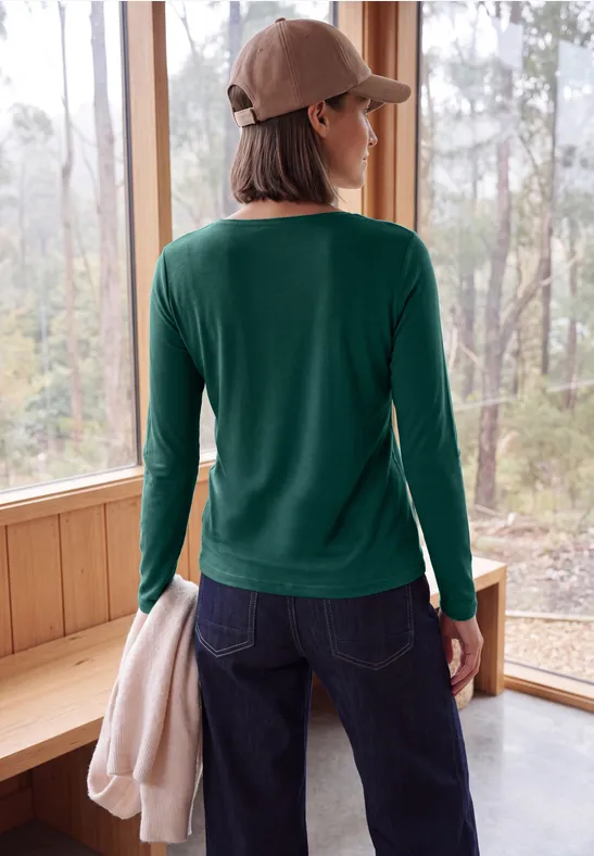 Frau mit grüner Bluse, Jeans und Cap, hält einen hellen Pullover vor einem großen Fenster im Wald.
