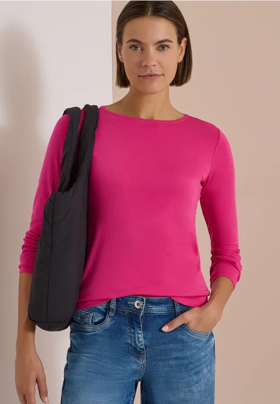 Frau mit pinkem Langarmshirt und Jeans trägt eine schwarze Tasche und steht vor hellem Hintergrund.