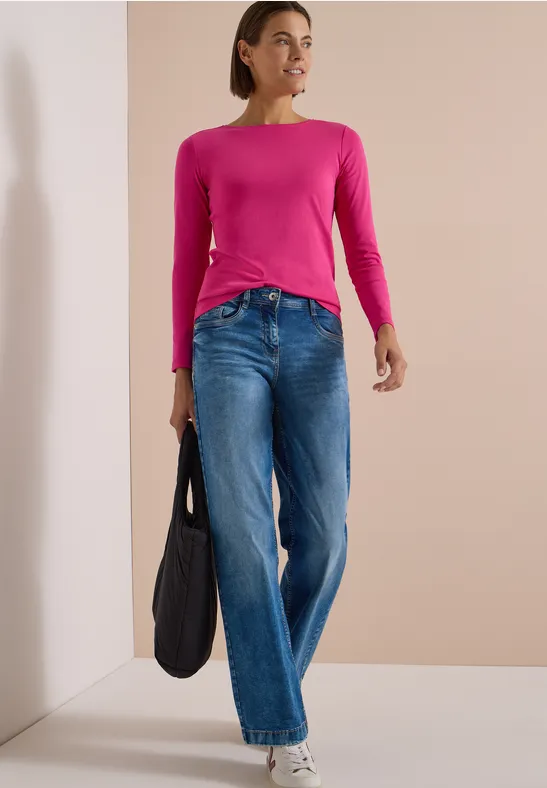 Frau in pinkem Langarmshirt und Jeans mit schwarzer Tasche in hellem Raum.