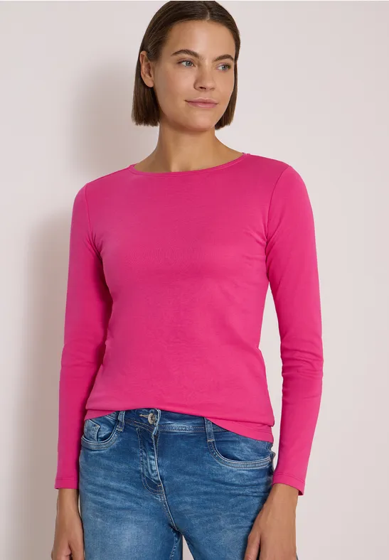 Eine Frau in pinkem Langarmshirt und blauen Jeans vor hellem Hintergrund.