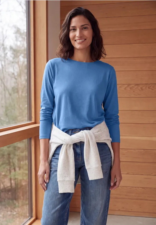 Eine lächelnde Frau in blauem Pullover und Jeans, mit weißem Sweatshirt um die Hüfte, in einem hölzernen Raum.