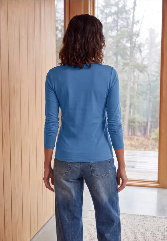 Person von hinten in blauem Oberteil und Jeans, stehend an Fenster in hellem Holzraum.