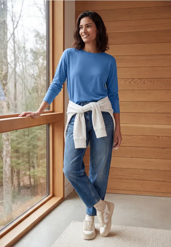 Frau in blauem Pullover und Jeans mit hellem Pullover um die Taille, lächelnd am Fenster