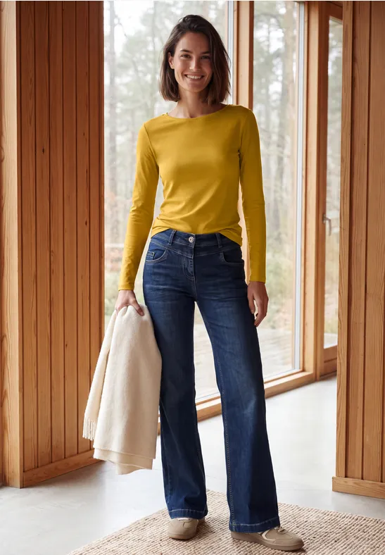 Frau im gelben Langarmshirt und blauen Jeans hält eine helle Jacke in modernem Raum.