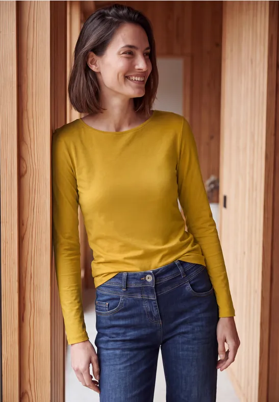 Lächelnde junge Frau in gelbem Langarmshirt und Jeans vor Holzwand.