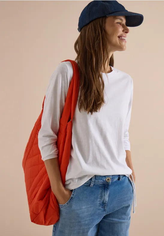 Femme souriante en t-shirt blanc, jean, sac rouge et casquette bleue, regardant sur le côté.