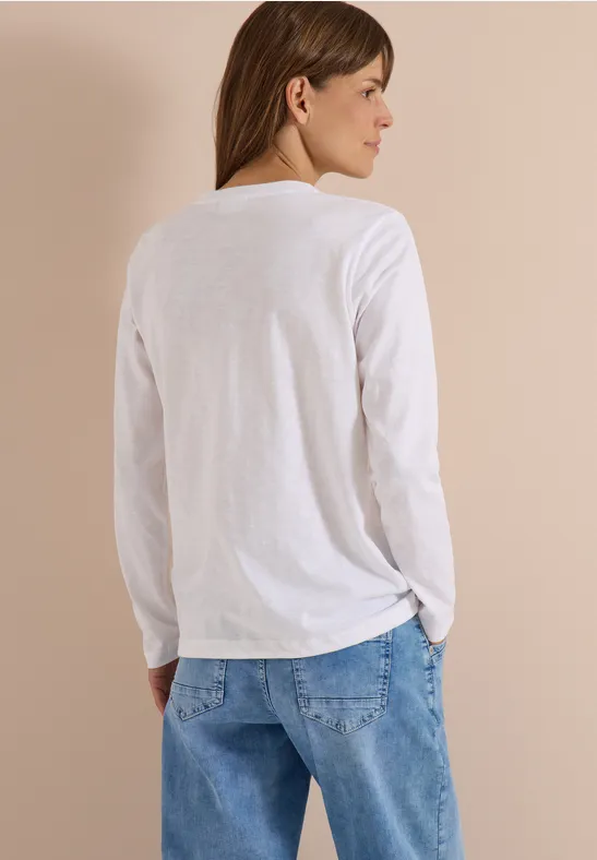 Une femme de dos porte un T-shirt blanc à manches longues et un jean bleu, regardant sur le côté.
