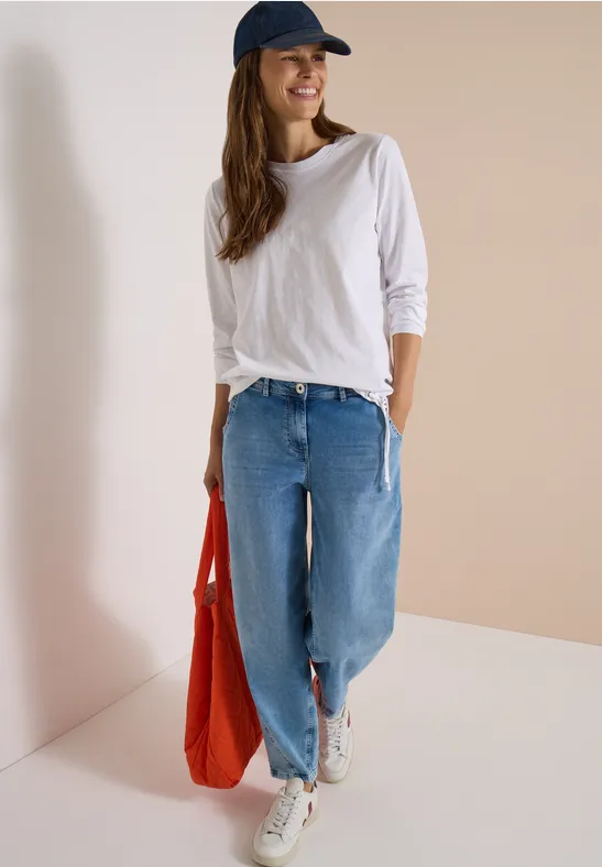 Une jeune femme en jean ample, t-shirt blanc et casquette bleue porte un sac rouge et baskets blanches.