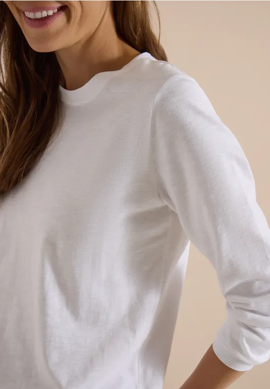 Femme portant un t-shirt à manches longues blanc, seul le buste et un sourire discret sont visibles.
