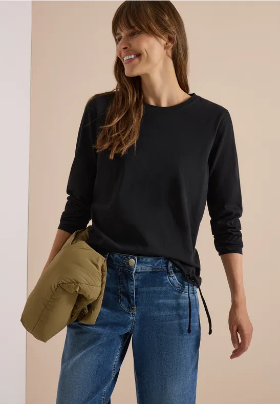Femme en t-shirt noir à manches longues et jean, tenant une veste kaki pliée, souriante.