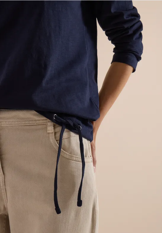 Une personne en pull bleu marine et pantalon crème à cordon, une main dans la poche.