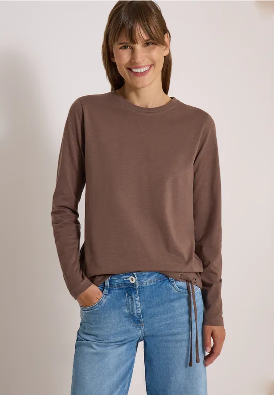 Femme souriante en t-shirt marron à manches longues et jean bleu, posant détendue les mains dans les poches.