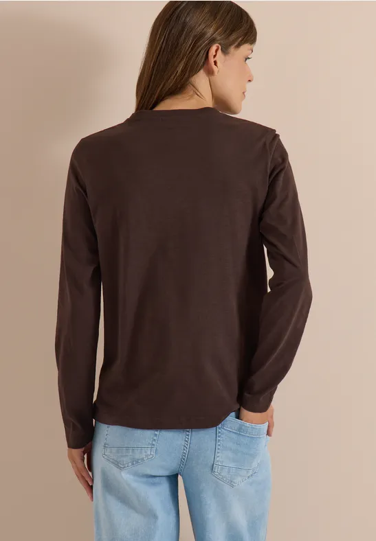 Rückansicht einer Frau in braunem Langarmshirt und hellblauen Jeans vor schlichtem Hintergrund.