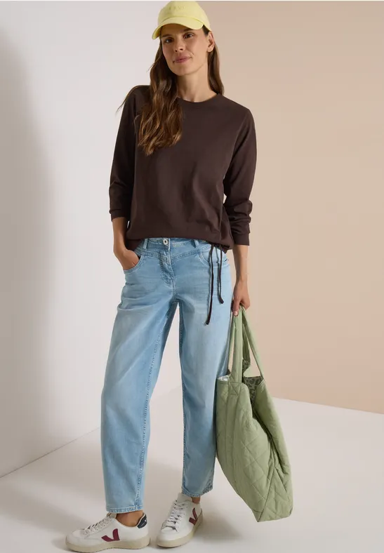 Eine junge Frau in lässigem Outfit: brauner Pullover, helle Jeans, weiße Sneaker, gelbe Mütze und große grüne Tasche.