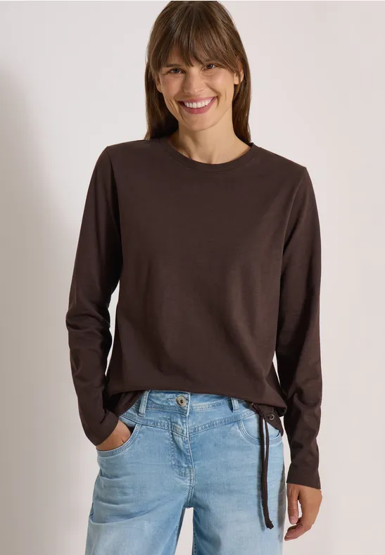 Eine lächelnde Frau trägt ein braunes Langarmshirt mit seitlicher Schleife und hellblaue Jeans.