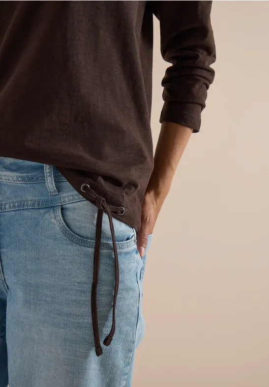 Teilansicht einer Person in braunem Langarmshirt und hellblauen Jeans mit seitlicher Schnürung.
