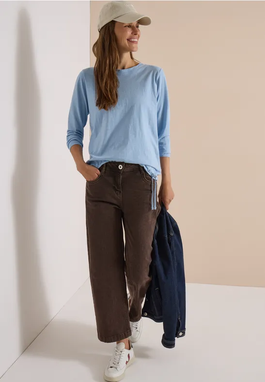 Femme en chemise bleu clair, pantalon large marron et casquette blanche appuyée contre un mur, sourire aux lèvres.