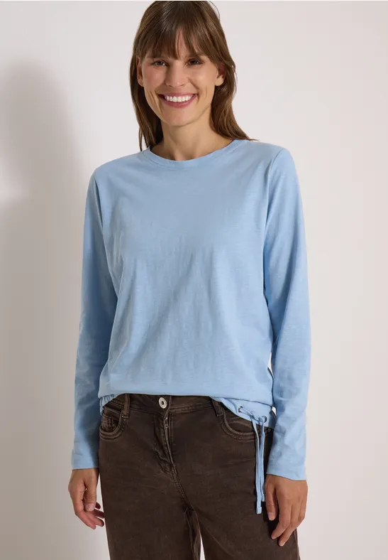 Une femme souriante en haut à manches longues bleu clair et pantalon en velours brun, fond blanc.