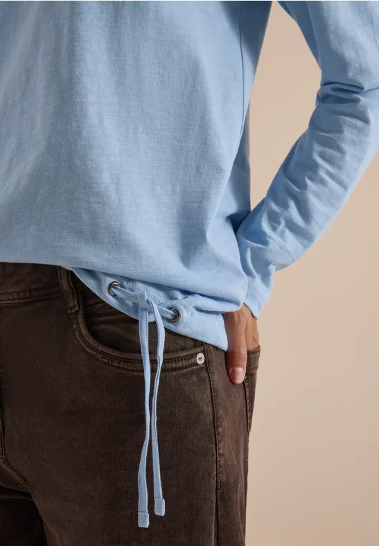 Pull bleu clair avec cordon de serrage, pantalon marron, main dans la poche sur fond beige.