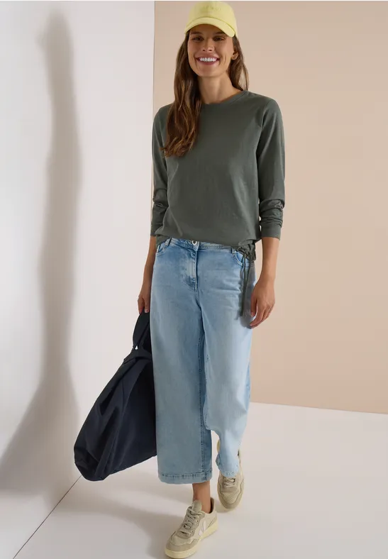 Femme en t-shirt vert à manches longues, jean bleu clair, casquette jaune, baskets et sac bleu foncé.