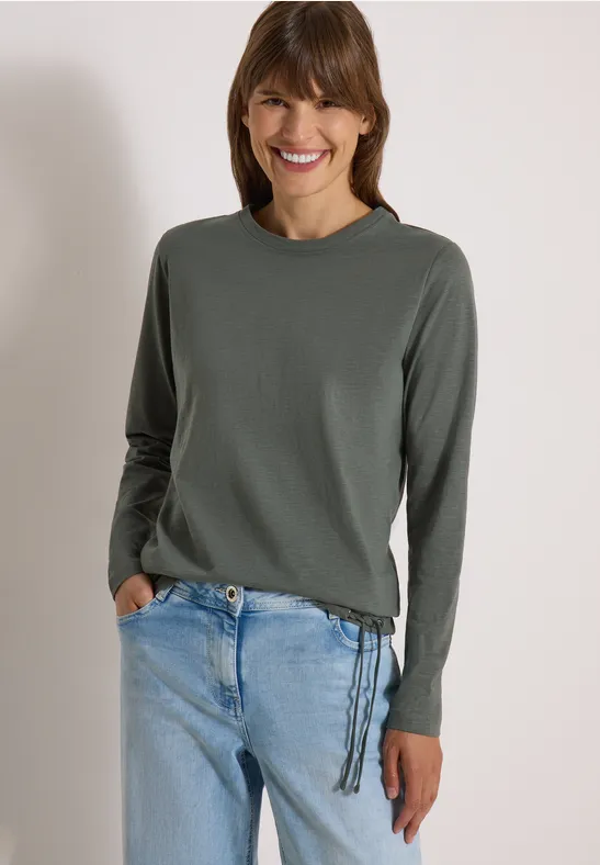Femme souriante en pull gris à lacets sur le côté et jean bleu clair.