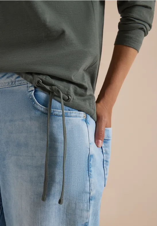 Gros plan d’une personne en sweat à capuche vert à cordons et jean bleu clair, une main dans la poche.