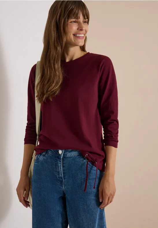 Une femme souriante porte un haut bordeaux à manches longues avec cordon de serrage et un jean bleu.