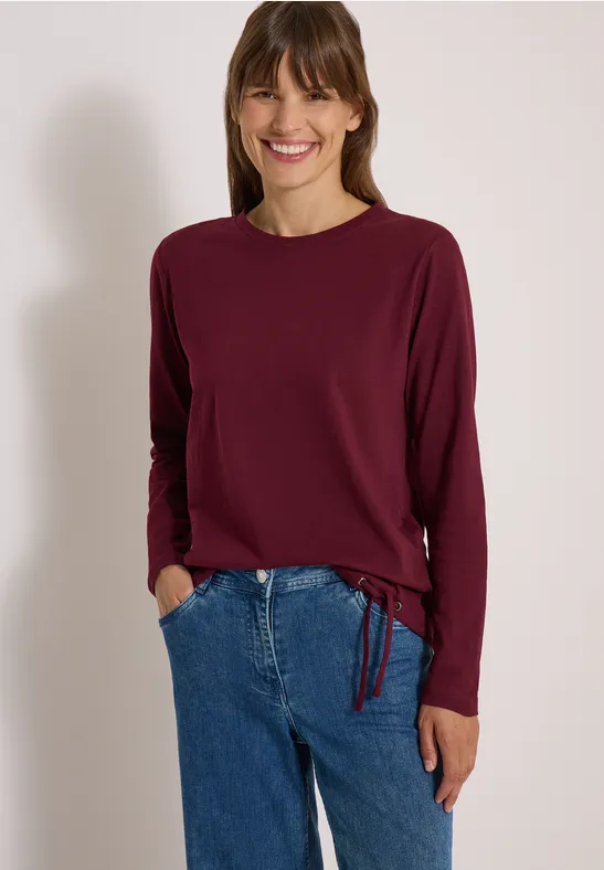 Femme souriante en haut à manches longues bordeaux et jean bleu devant un fond neutre.